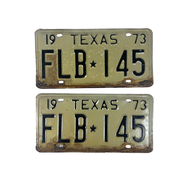 Pair Of 1973 Texas License Plates FLB 145 Vintage White w Black Embossed Letters. Two vintage 1973 Texas license plates with the plate number FLB 145, featuring a faded olive-green background and black lettering, displayed on a white textured fabric background.