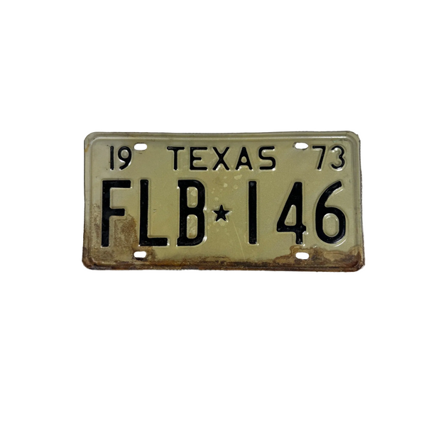 Vintage 1973 Texas License Plate FLB 146 Black Embossed Lettering On White Metal. A vintage 1973 Texas license plate reading FLB 146, featuring a faded tan background with black lettering and weathered rust-like discoloration along the bottom edge, displayed on a textured white surface.