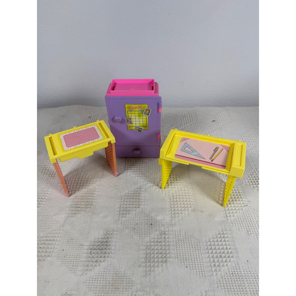 Barbie Stacie 1993 Locker and School Desk Playset Vintage Doll Furniture. A vintage toy playset featuring a purple and pink safe in the center, flanked by two small yellow desks with pink and red surfaces, displayed on a textured light gray surface against a neutral gray wall.