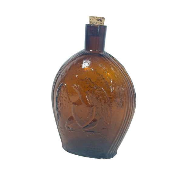 Vintage Clevenger Amber Glass Bottle w Embossed Eagle and Grapes Pattern 1950s. An antique amber glass perfume bottle with a cork stopper, featuring an embossed figure on the front, set against a light blue background and white textured fabric.