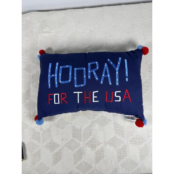Hooray For The USA Decorative Pillow w Patriotic Pom Pom Tassels Americana. A rectangular navy blue throw pillow with 'HOORAY! FOR THE USA' embroidered in red, white, and blue, resting on a textured white blanket.