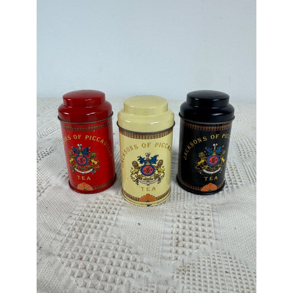 Jacksons Of Piccadilly Tea Tin Set Of 3 Metal Containers Red Yellow Black Tins, Three vintage metal tea tins with screw-on lids, displayed side-by-side on a white textured crochet blanket. The left tin is red, the middle tin is cream/beige, and the right tin is black.