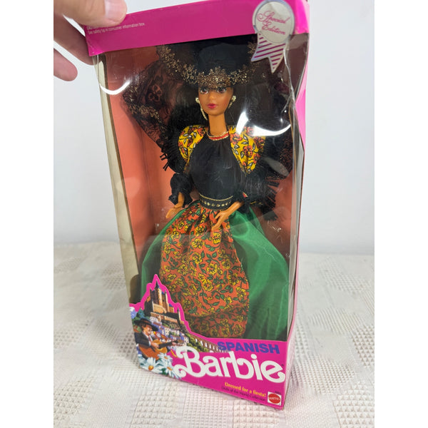 A vintage Barbie doll in its original pink packaging, held by a hand. The doll is labeled 'Spanish Barbie' and features a black dress with yellow patterned details and a green skirt. Spanish Barbie Dolls Of The World Collection 1991 Special Edition 4963 NIB 