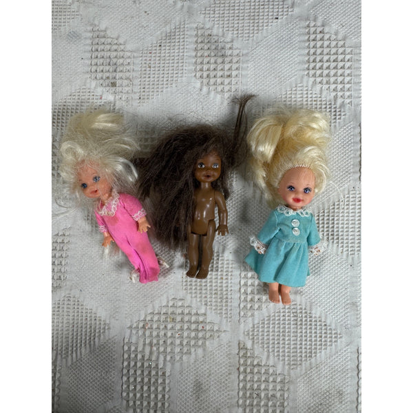 Set of 3 Vintage 1990s Kelly and Friends Bedtime Baby Dolls Bottle Mouth Barbie. Three vintage plastic dolls with messy hair lying on a white textured fabric background. One doll has white hair and a pink outfit, one has dark brown hair and no outfit, and one has blonde hair and a light blue dress.