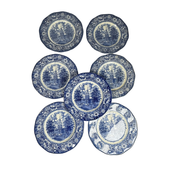Enoch Wedgewood Liberty Blue 10" Ironstone Plates Set 7 Liberty Hall 1970s. Six blue and white ceramic plates with a classic transfer design of trees and architectural scenes, arranged on a textured cream tablecloth.