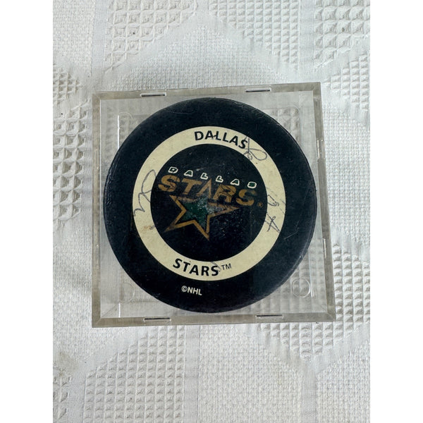 Dallas Stars NHL Official Game Puck w Signatures In Acrylic Case Autographed. A Dallas Stars hockey puck inside a clear plastic protective case, placed on a textured white paper towel background. The puck features the Dallas Stars logo with gold and white details on a black background, with autograph markings visible.
