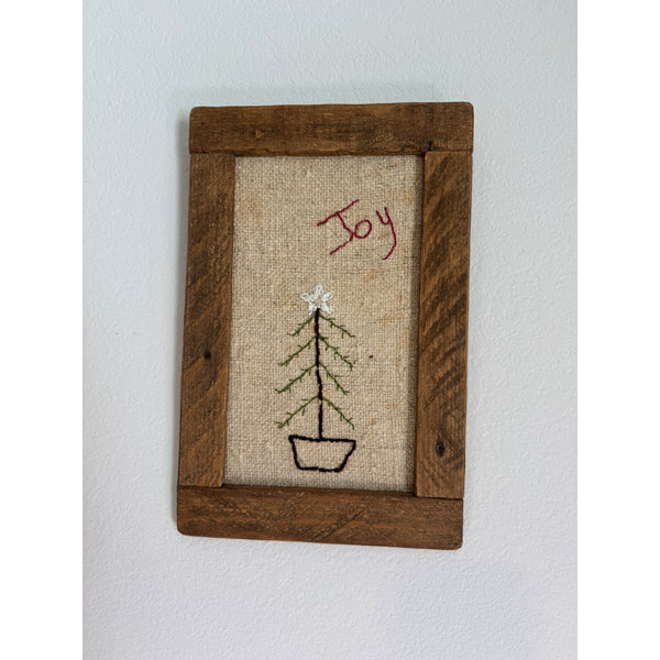 Hand Embroidered Rustic Joy Christmas Tree Wall Art In Wooden Frame Farmhouse. A rustic framed wall hanging with a dark brown wooden frame, featuring a burlap-textured center with a simple embroidered Christmas tree and the word 'Joy' stitched in red.
