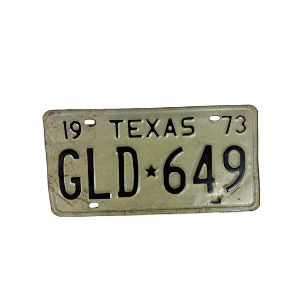 Vintage 1973 Texas License Plate GLD 649 White w Black Embossed Lettering Metal. A vintage 1973 Texas license plate with the plate number GLD 649, featuring a light beige background and black lettering, displayed on a textured white surface.