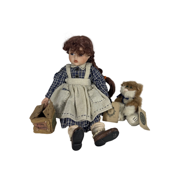 Boyds Bears Yesterdays Child Porcelain Doll "Jamie" With Plush Cat & Kitten Box. A vintage-style doll with brown hair sits on a white quilted surface against a light gray wall, holding a small brown box. The doll wears a blue-and-white plaid shirt and apron.