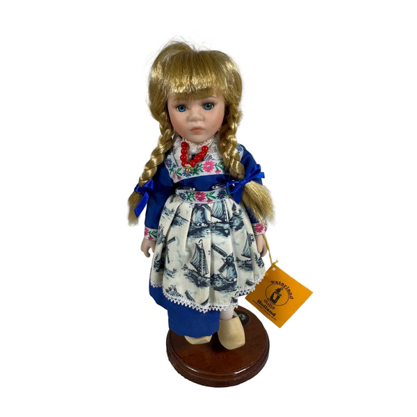 Vintage Waterland Holland Doll Porcelain 1990s Traditional Costume with Stand. A vintage German porcelain doll with blonde braided hair, wearing a blue and white patterned dress, standing on a wooden base with a yellow brand tag attached.