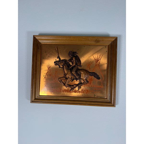 Vintage John Louw Copper Embossed Wall Art Of Native American On Horse 1970s. A framed vintage wall art piece featuring a dark metal cutout of a Native American rider on a horse against a warm gold-toned background, mounted on a light blue-gray wall.