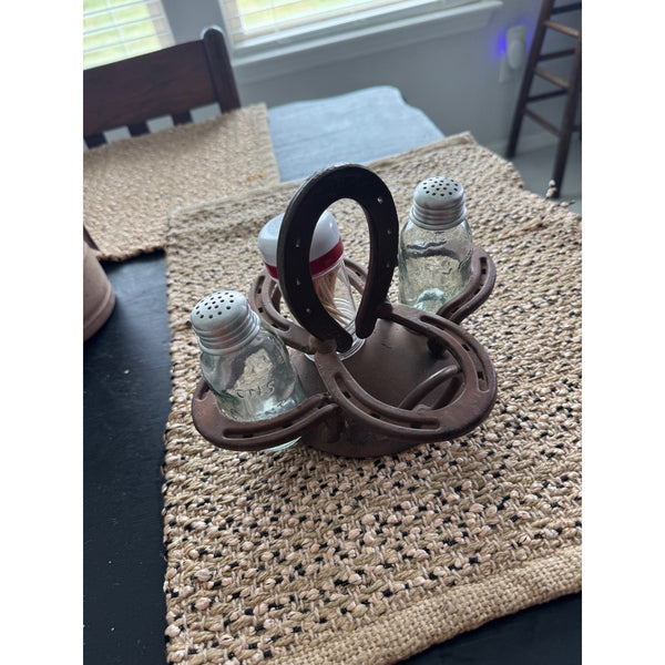 Rustic Horseshoe Salt & Pepper Shaker Spice Holder Tabletop Caddy Metal Western. A rustic metal horseshoe-shaped salt and pepper shaker holder with three glass shakers, displayed on a woven placemat on a dark table.