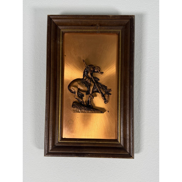 Native American Warrior Embossed Copper Wall Art With Wooden Frame Vintage 1970s. A framed decorative wall piece featuring a bronze-toned sculpted figure of a rider on horseback against a warm gold background, enclosed in a dark brown wooden frame and mounted on a light gray wall.