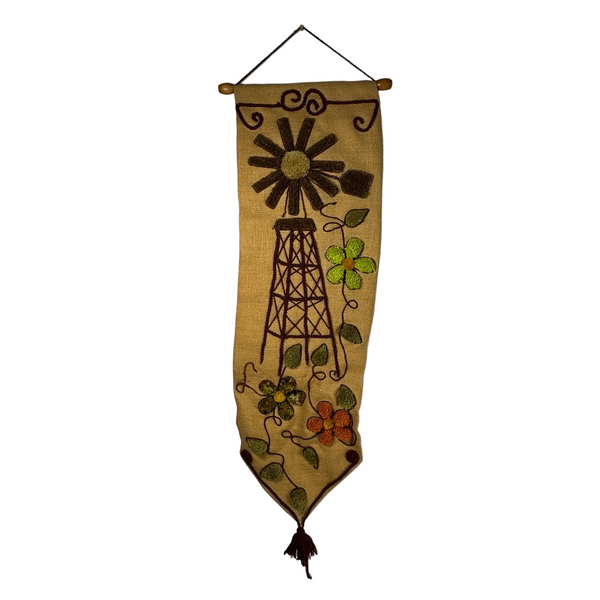 Handcrafted Burlap Yarn Wall Hanging w Windmill Floral Design 1970s Vintage. A vertical woven wall hanging banner made from tan burlap, featuring a dark brown oil derrick and sun motif with green and orange flowers, hanging against a plain light gray wall.