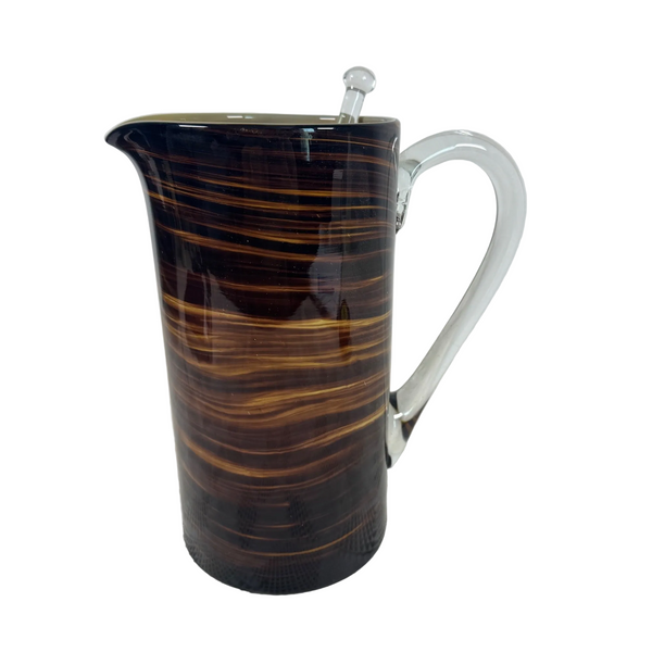 Handblown Glass Tortoise Shell Pitcher w Swirled Amber Brown Pattern And Stirrer. A glass pitcher with a glossy dark brown and amber swirl pattern and a clear glass handle, shown on a white textured surface against a light gray background.