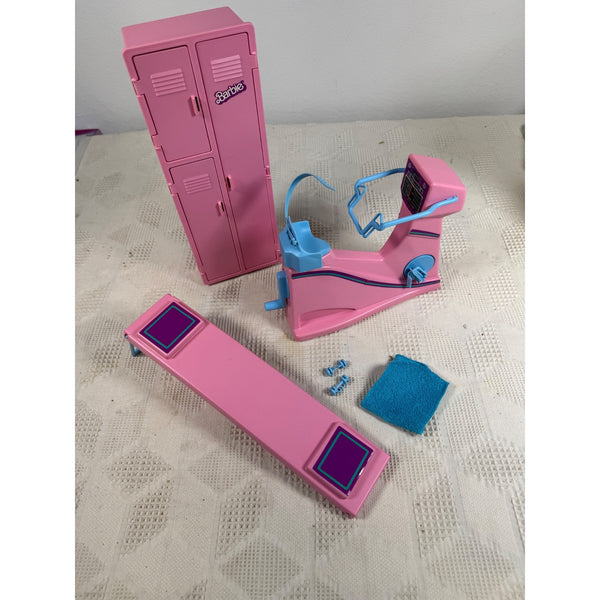 1984 Barbie Workout Center Pink Locker Bike Bench Weights Towel Partial Set. A pink toy locker and exercise bike set with blue accents, shown on a textured off-white surface. The set includes a pink locker with a small 'Snoopy' badge, an exercise bike with blue handlebars and pedals, a pink platform with purple pads, and a small blue cloth.