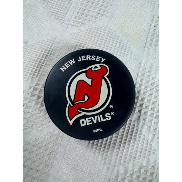 New Jersey Devils Official Licensed NHL Hockey Puck by InGlasCo Made In Slovakia. A circular black New Jersey Devils hockey puck badge with the team logo, resting on a white textured mesh surface.