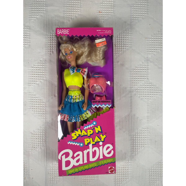 A boxed Barbie doll with long blonde hair, wearing a yellow top and blue skirt, shown in its pink retail packaging on a textured white surface. The box reads 'Barbie' and 'Snap 'n Play'.
