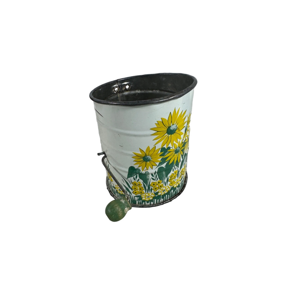 Vintage Metal Flour Sifter With Hand Crank and Sunflower Design Retro Kitchen. A vintage metal bucket with a white body decorated with yellow sunflowers and green leaves, featuring a dark metal rim and a small green handle, resting on a white textured surface.