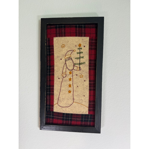 Framed Santa Embroidery On Plaid Background Rustic Black Wooden Frame Farmhouse. A framed handmade folk art piece featuring a stitched snowman holding a pine tree, set against a red and black plaid background and enclosed in a black frame, mounted on a white wall.