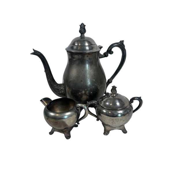Vintage Ornate Silver Plated Tea Set Includes Teapot Sugar Bowl and Creamer 1940s. A vintage three-piece silver tea or coffee set including a teapot, a small sugar bowl, and a small creamer, displayed on a textured white tablecloth against a plain light gray wall.