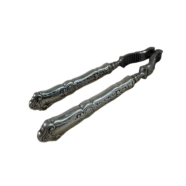 Vintage Sterling Silver Nutcracker With Intricate Handle Design Antique c1920s. An ornate silver-toned metal tongs or clamps with decorative engraved handles, placed on a textured off-white woven fabric surface.