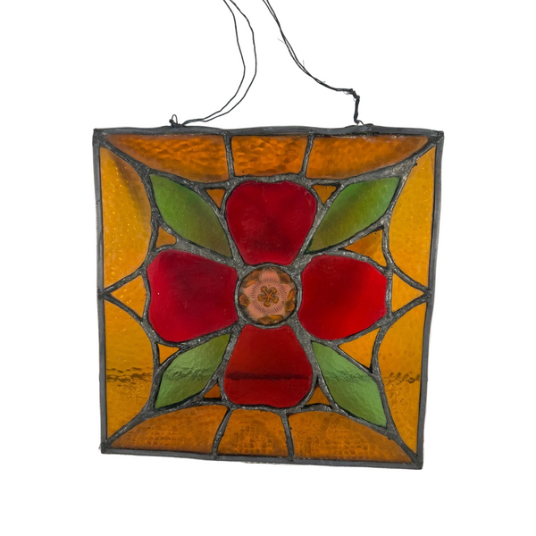 Vintage 12 Inch Stained Glass Panel Floral Design Leaded Stained Glass Wall Art. A square stained glass panel featuring a central red flower with green leaves and a gold-toned center, surrounded by amber and orange sections, suspended by black wires against a plain wall.