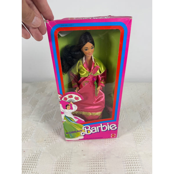 A boxed Barbie doll with a hand holding the top of the package, shown against a light neutral background. The doll is dressed in a pink and green traditional-style outfit and is visible through a clear window on the front of the box. Korean Barbie Doll Mattel 1990s Dolls Of The World Collection 4929 NIB