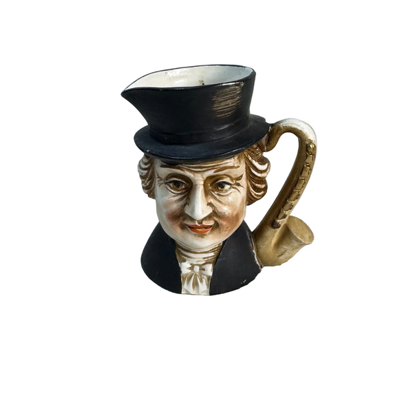 Vintage Ceramic Toby Face Character Creamer Top Hat and Pipe Handle Lego Japan. A ceramic pitcher shaped like a man's face wearing a black top hat and dark coat, with a gold-toned handle and white shirt details, set on a textured white surface against a light gray wall.