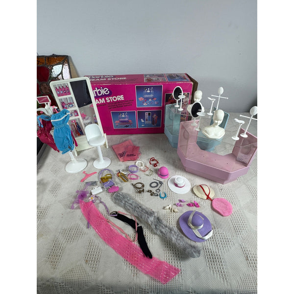 Barbie Dream Store Fashion Department Playset Accessories and Box Vintage 1980s. A Barbie doll accessories set arranged on a light patterned tablecloth, including a pink display stand, white chairs, jewelry, a blue dress, and a pink Barbie store box in the background.