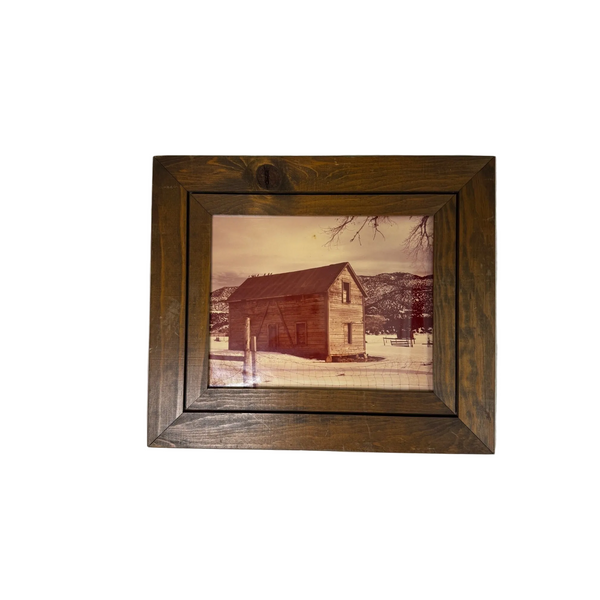 Rustic Wooden Framed Print Of Abandoned Cabin Winter Scene w Mountains and Fence. A rustic dark brown wooden picture frame containing a vintage-style photograph of a wooden barn in a snowy landscape, mounted on a light gray textured wall.